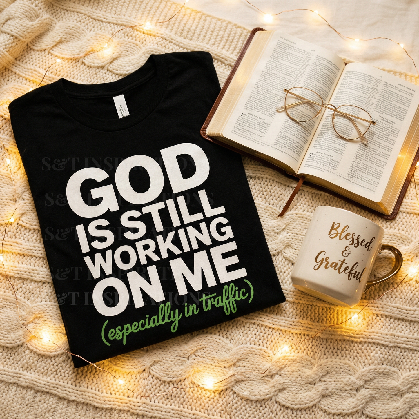 Black t-shirt with 'GOD IS STILL WORKING ON ME (especially in traffic)' text, mug, and open book on textured surface with lights.