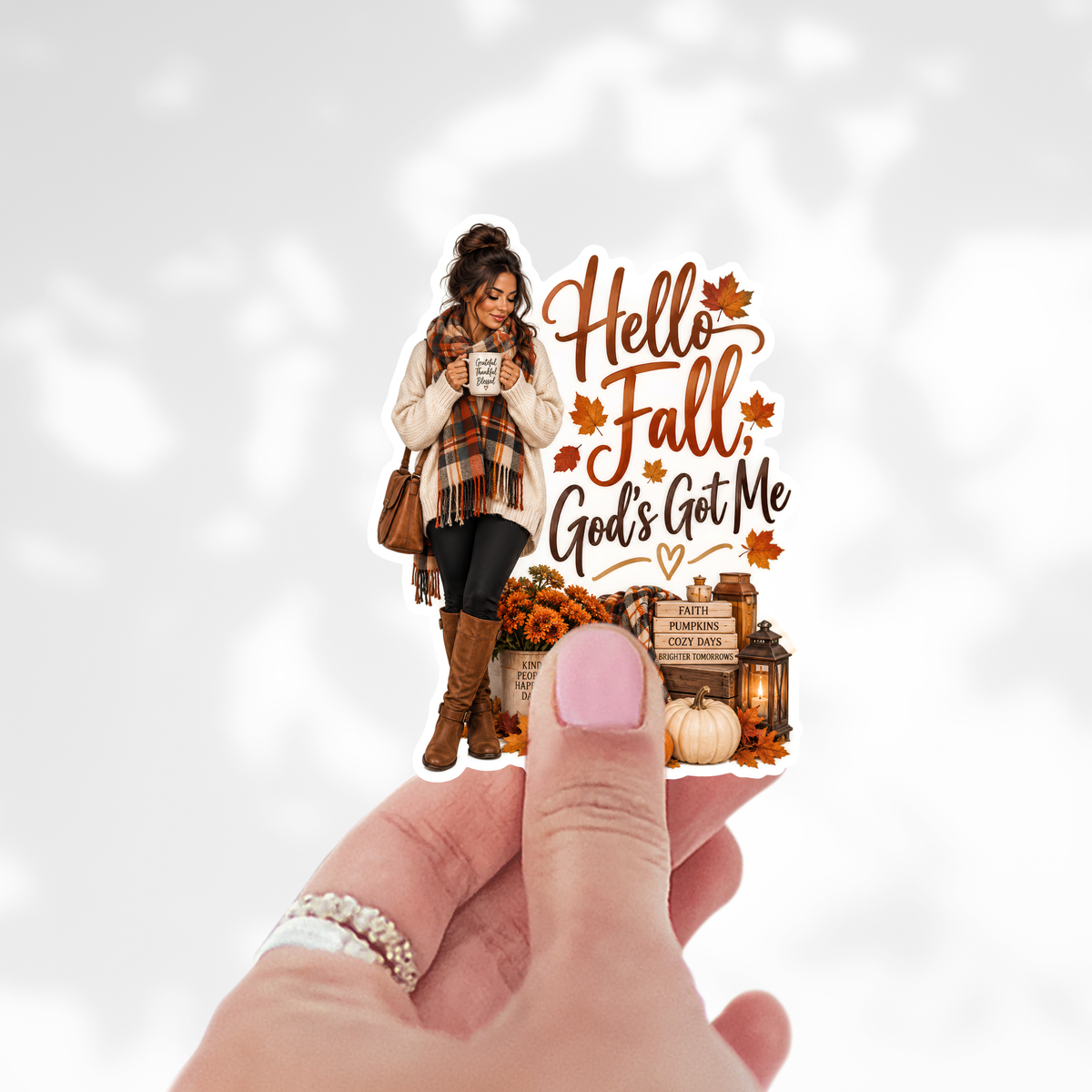 A hand with pink nail polish holds a die-cut sticker featuring an autumn-themed illustration of a woman in fall clothing next to the text 'Hello Fall God's Got Me' with pumpkins, leaves, and lanterns, against a soft white background with faint leaf shadows.