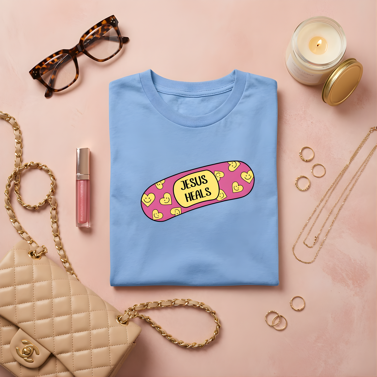 Light blue t-shirt with 'Jesus Heals' design on a pink surface with accessories.