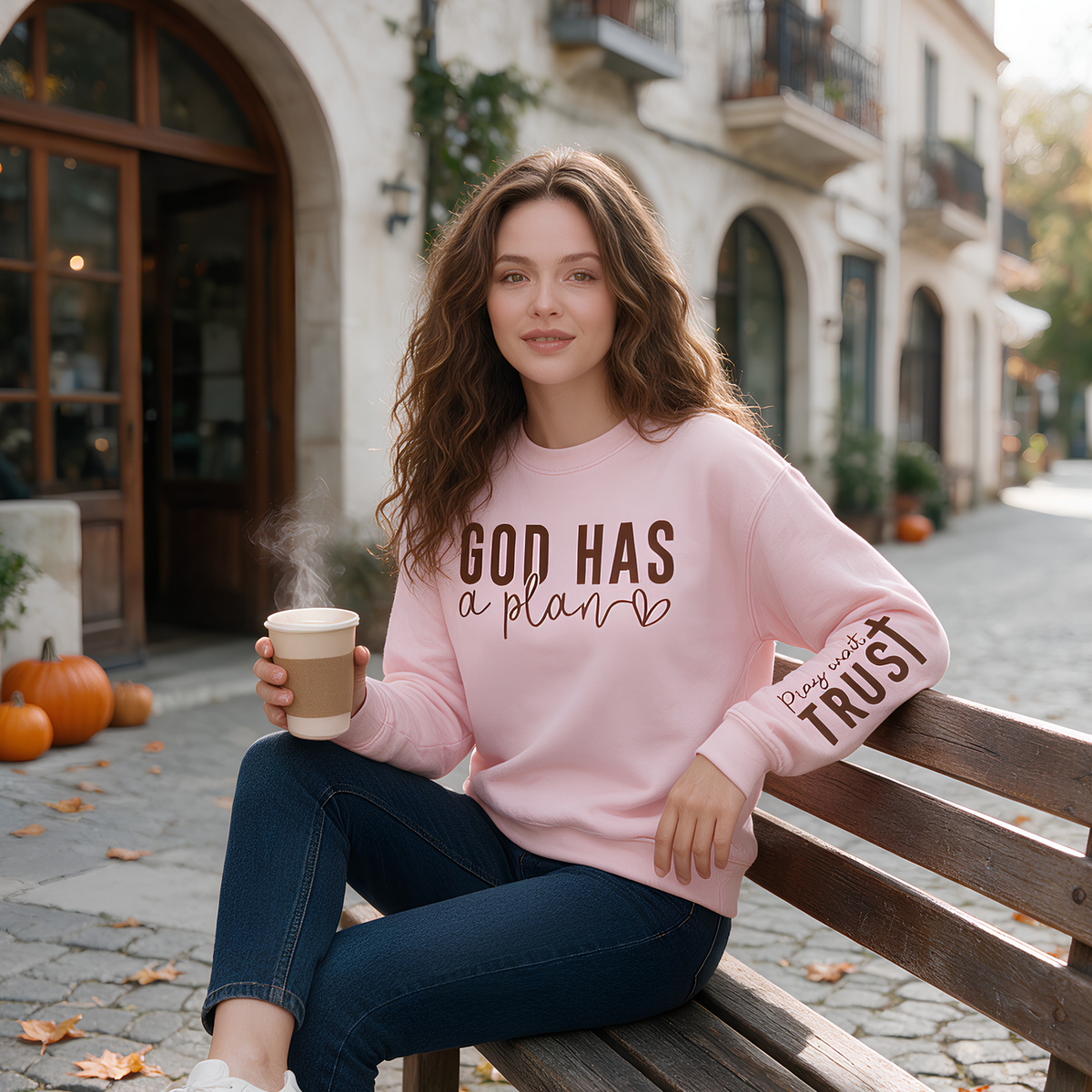 A woman sits on a wooden bench outdoors in front of a cafe, wearing a light pink crewneck sweatshirt with the text 'GOD HAS a plan' on the chest and 'Please believe TRUST' on the sleeve, paired with dark blue jeans and holding a coffee cup.