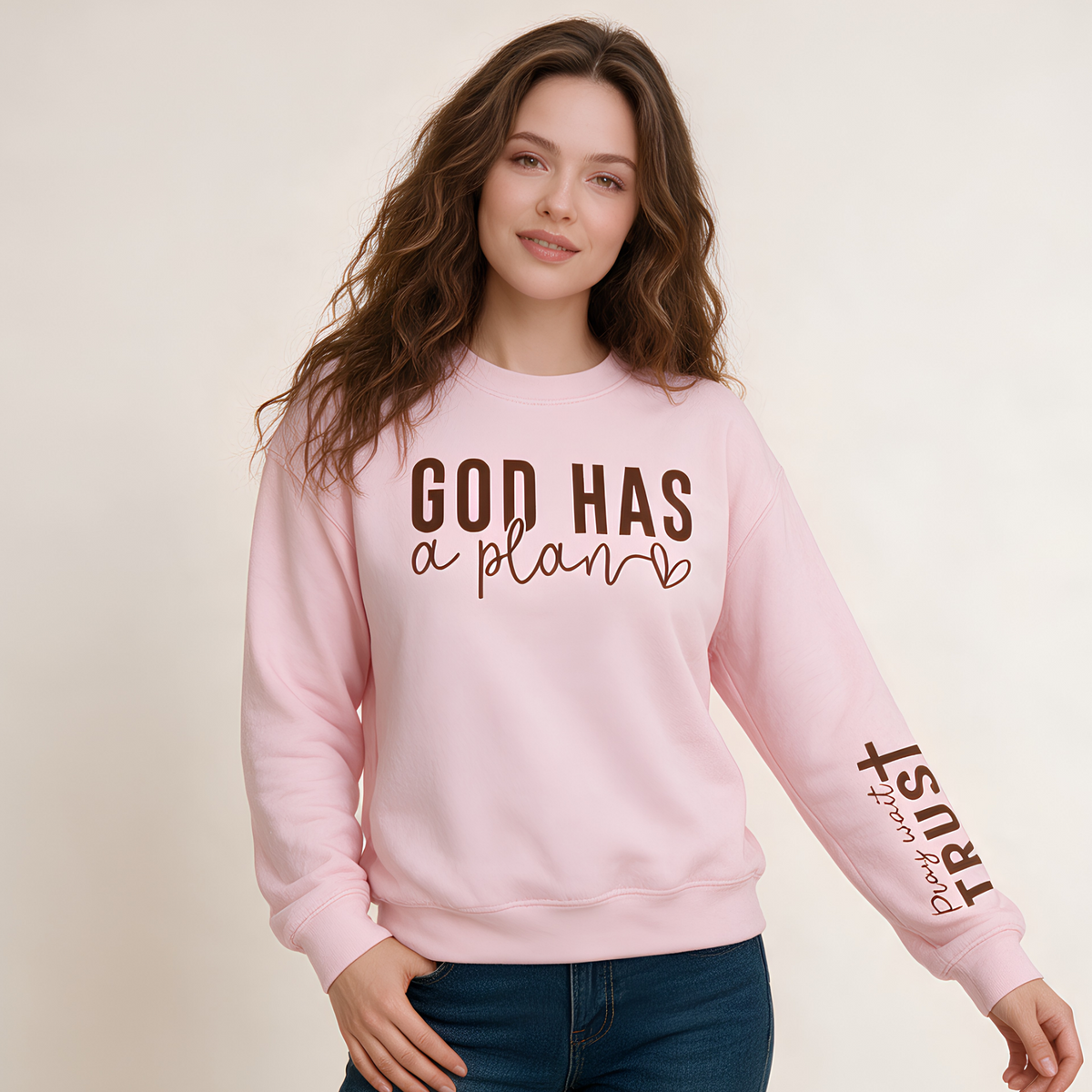 A woman modeling a light pink crewneck sweatshirt with brown text that reads 'GOD HAS a plan' across the chest and 'trust PINK' printed on the sleeve, paired with blue jeans against a plain light beige background.