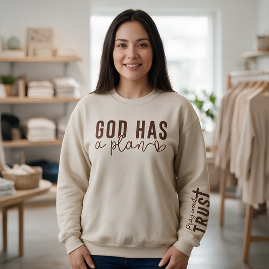 A woman wearing a cream-colored crewneck sweatshirt with brown text that reads 'GOD HAS a plan' on the front and 'May worth TRUST' on the sleeve, standing in a softly blurred boutique setting.