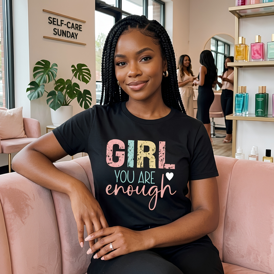 A woman with braided hair sits on a pink couch in a salon-like interior, wearing a black graphic T-shirt that reads “GIRL YOU ARE enough” in pastel and white lettering. Other salon chairs, shelves with bottles, and a sign reading “SELF-CARE SUNDAY” are visible in the background.