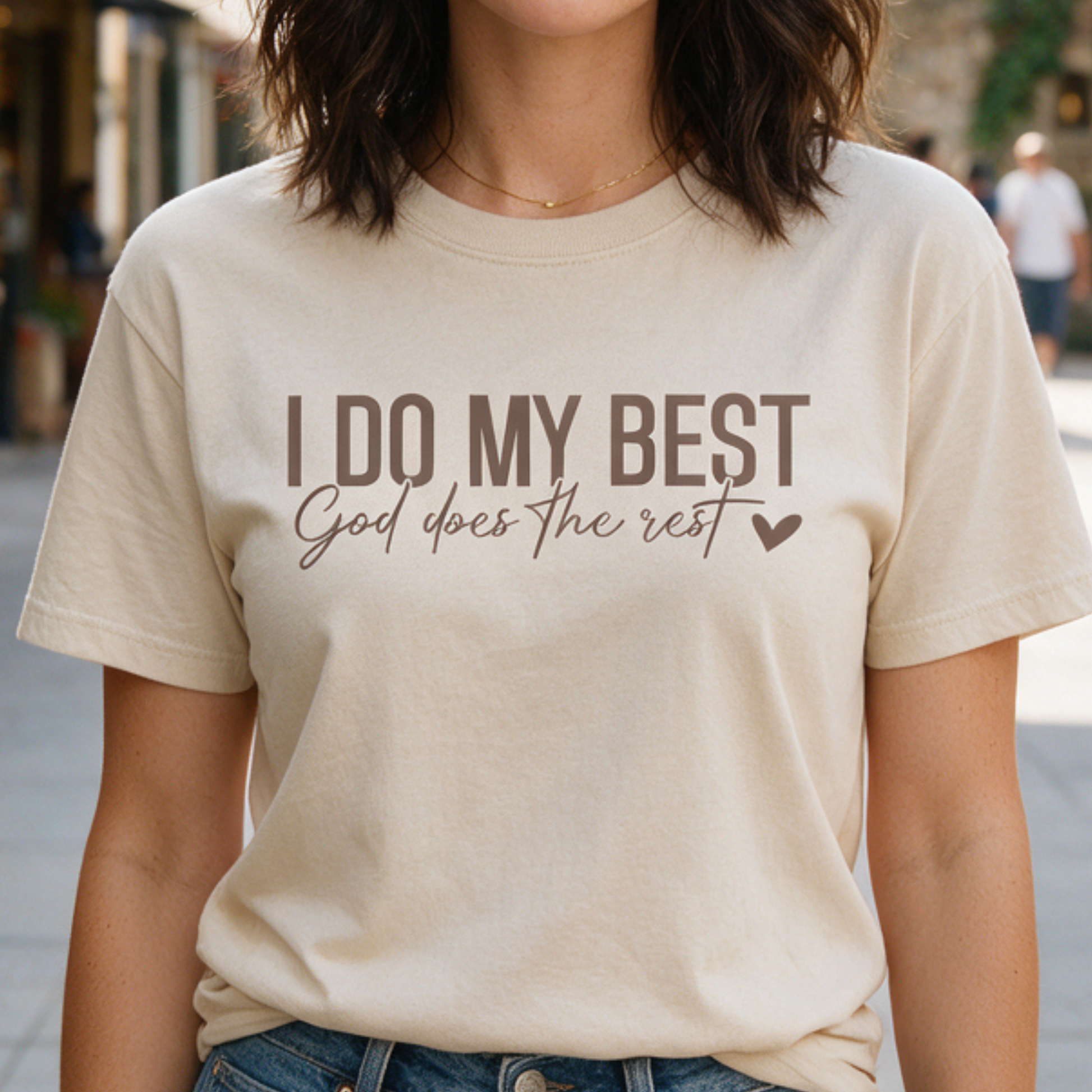 A woman wearing a cream-colored t-shirt with the text 'I DO MY BEST God does the rest' printed in brown, shown in an outdoor setting.